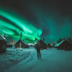 En person ser på norrskenets magi över snötäckta stugor. Aurora borealis, som latin kallar det, lyser upp natthimlen.