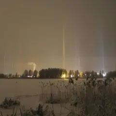 Vintermörker med lysande ljuspelare över en snötäckt sjö. En fabrik släpper ut rök i bakgrunden.