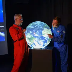 Två män i rymddräkter pekar på en lysande jordglob, som representerar vår planet.