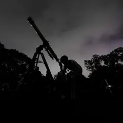 Mörk silhuett av en person som tittar i ett teleskop under en molnig natt. Träd och andra människor syns i bakgrunden, med svagt **stray light** i himlen.