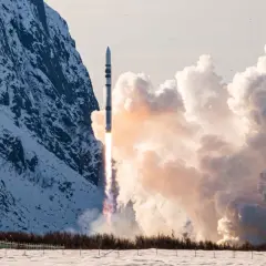 ISAR Aerospace raket skjuter upp från en snötäckt mark, med en snöig bergssida och moln i bakgrunden.