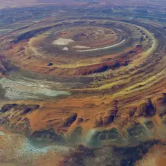 Ökenlandskap med ringformade formationer, som ett gigantiskt öga i Sahara. Färger av orange, gult och blått skapar ett surrealistiskt mönster.