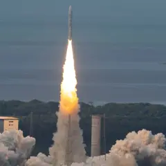 Ariane 5 skjuter upp från marken med en enorm eldsvåda och rök. Raketen stiger mot en blå himmel över ett grönt landskap och ett lugnt hav.
