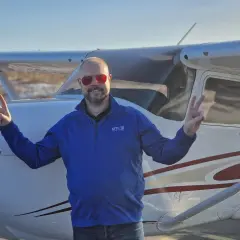Man in blue shirt and red sunglasses making peace signs in front of a small airplane.