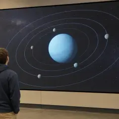 En person tittar på en stor skärm som visar uranus omloppsbana runt solen, med flera månar som kretsar kring planeten.