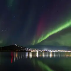 Kiruna norrsken lyser upp natthimlen över en stad vid en sjö. Gröna och lila ljusstrimmor dansar över stjärnorna.