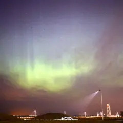 Norrsken lyser upp natthimlen, kanske tack vare solstormar idag. Grönaktiga ljusstrimmor dansar över ett landskap med kullar och gatlyktor.