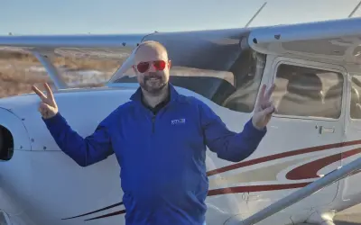 Man in blue shirt and red sunglasses making peace signs in front of a small airplane.