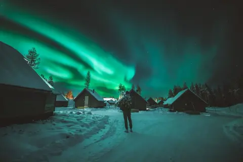 En person ser på norrskenets magi över snötäckta stugor. Aurora borealis, som latin kallar det, lyser upp natthimlen.