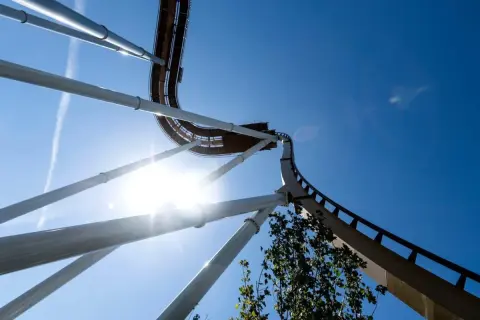 En berg-och-dalbana som svänger upp mot en klarblå himmel, solen lyser starkt och skapar en bländande effekt. Känslan av g krafter är påtaglig.