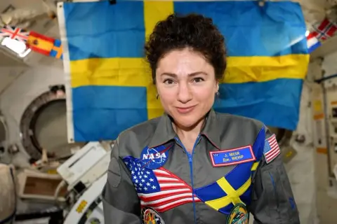 Astronaut med svensk flagga bakom sig. Hon bär en dräkt med både amerikanska och svenska symboler, vilket visar att svenskar i rymden är en del av internationellt samarbete.