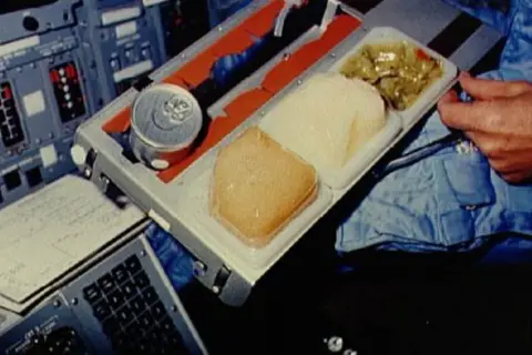 En astronaut äter rymdmat från en bricka. Maten ser ut som en burk, en gelé och en grönsaksrätt.