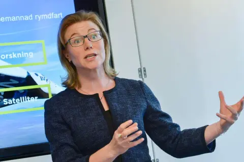 Karin Nilsdotter, med glasögon, gestikulerar vid en skärm som visar "Forskning" och "Satelliter" i samband med rymdfart.