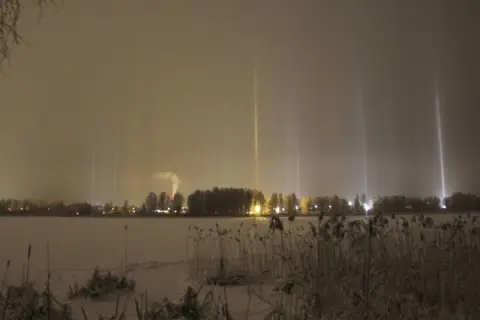Vintermörker med lysande ljuspelare över en snötäckt sjö. En fabrik släpper ut rök i bakgrunden.