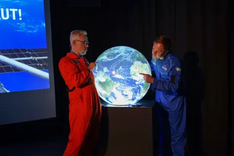 Två män i rymddräkter pekar på en lysande jordglob, som representerar vår planet.