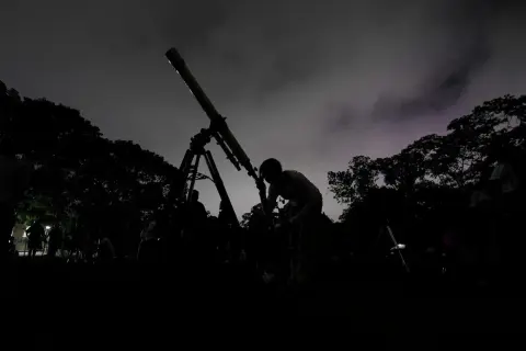 Mörk silhuett av en person som tittar i ett teleskop under en molnig natt. Träd och andra människor syns i bakgrunden, med svagt **stray light** i himlen.