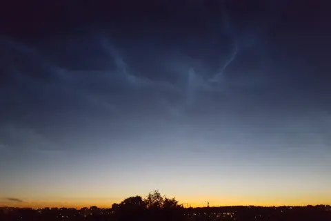Mörka moln med ljusa strimmor, nattlysande moln, över en stad i skymningen.