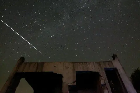 En ljus meteor drar en strimma över en stjärnklar natthimmel, ovanför ruinerna av en byggnad.