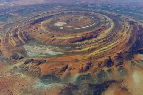 Ökenlandskap med ringformade formationer, som ett gigantiskt öga i Sahara. Färger av orange, gult och blått skapar ett surrealistiskt mönster.