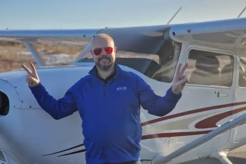 Man in blue shirt and red sunglasses making peace signs in front of a small airplane.