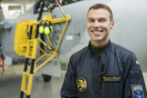 Marcus Wandt, Gripen testpilot, ler glatt framför ett stridsflygplan.