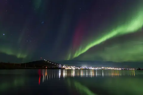 Kiruna norrsken lyser upp natthimlen över en stad vid en sjö. Gröna och lila ljusstrimmor dansar över stjärnorna.