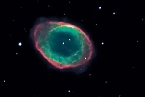 Den kosmiska ringnebulosan lyser i grönt och rosa mot en mörk stjärnhimmel.