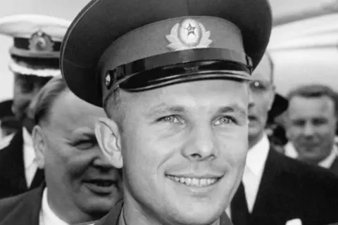 Juri Gagarin, den första människan i rymden, ler mot kameran. Han bär en rymdkeps med en sovjetisk symbol.
