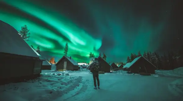 En person ser på norrskenets magi över snötäckta stugor. Aurora borealis, som latin kallar det, lyser upp natthimlen.