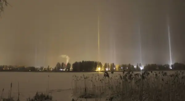 Vintermörker med lysande ljuspelare över en snötäckt sjö. En fabrik släpper ut rök i bakgrunden.
