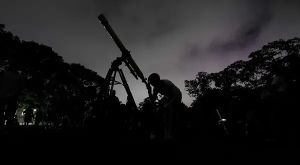Mörk silhuett av en person som tittar i ett teleskop under en molnig natt. Träd och andra människor syns i bakgrunden, med svagt **stray light** i himlen.