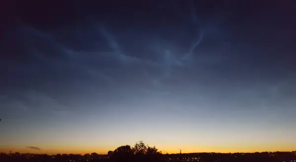 Mörka moln med ljusa strimmor, nattlysande moln, över en stad i skymningen.