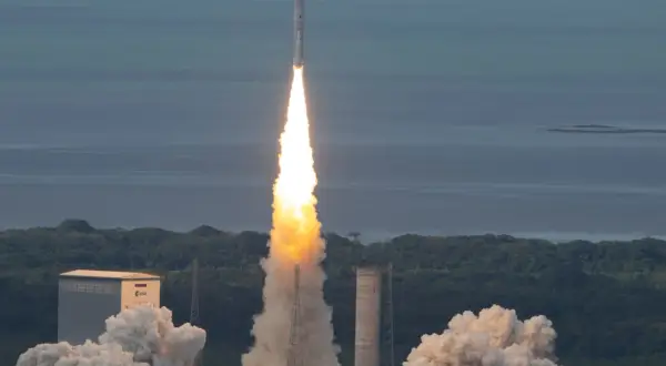 Ariane 5 skjuter upp från marken med en enorm eldsvåda och rök. Raketen stiger mot en blå himmel över ett grönt landskap och ett lugnt hav.