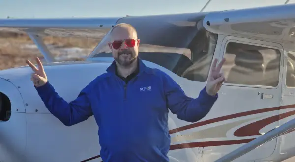 Man in blue shirt and red sunglasses making peace signs in front of a small airplane.