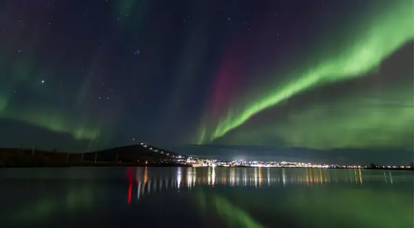 Kiruna norrsken lyser upp natthimlen över en stad vid en sjö. Gröna och lila ljusstrimmor dansar över stjärnorna.