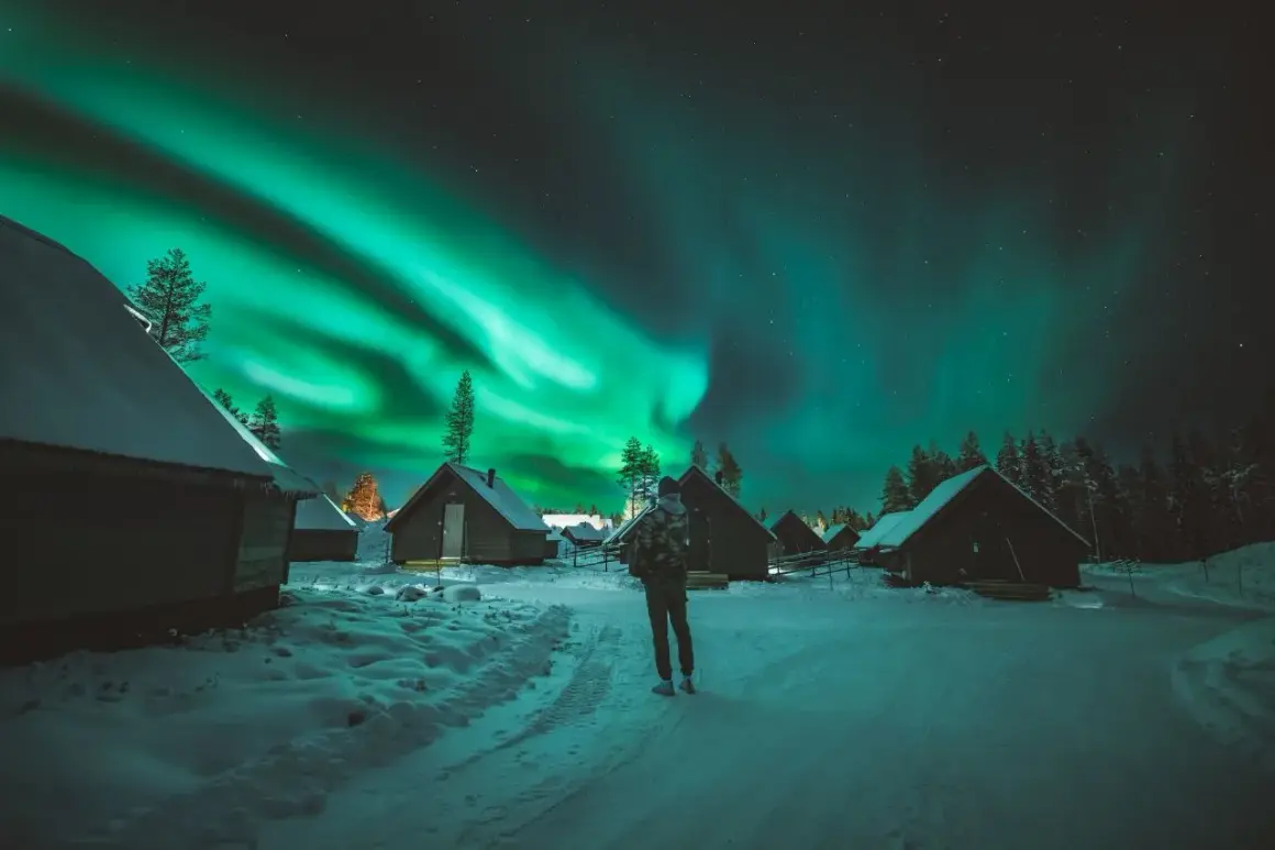 En person ser på norrskenets magi över snötäckta stugor. Aurora borealis, som latin kallar det, lyser upp natthimlen.