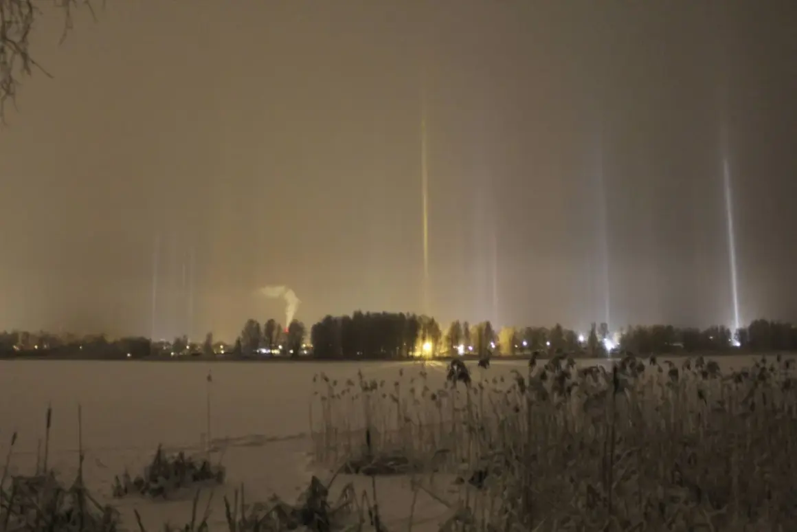 Vintermörker med lysande ljuspelare över en snötäckt sjö. En fabrik släpper ut rök i bakgrunden.