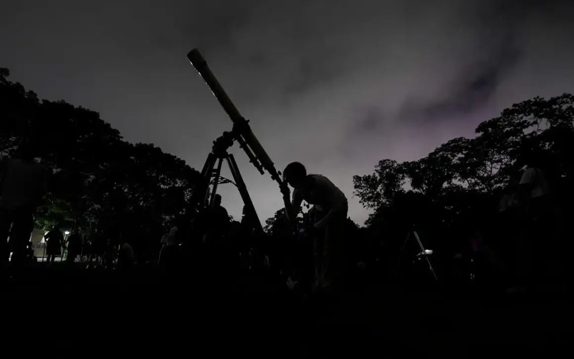 Mörk silhuett av en person som tittar i ett teleskop under en molnig natt. Träd och andra människor syns i bakgrunden, med svagt **stray light** i himlen.