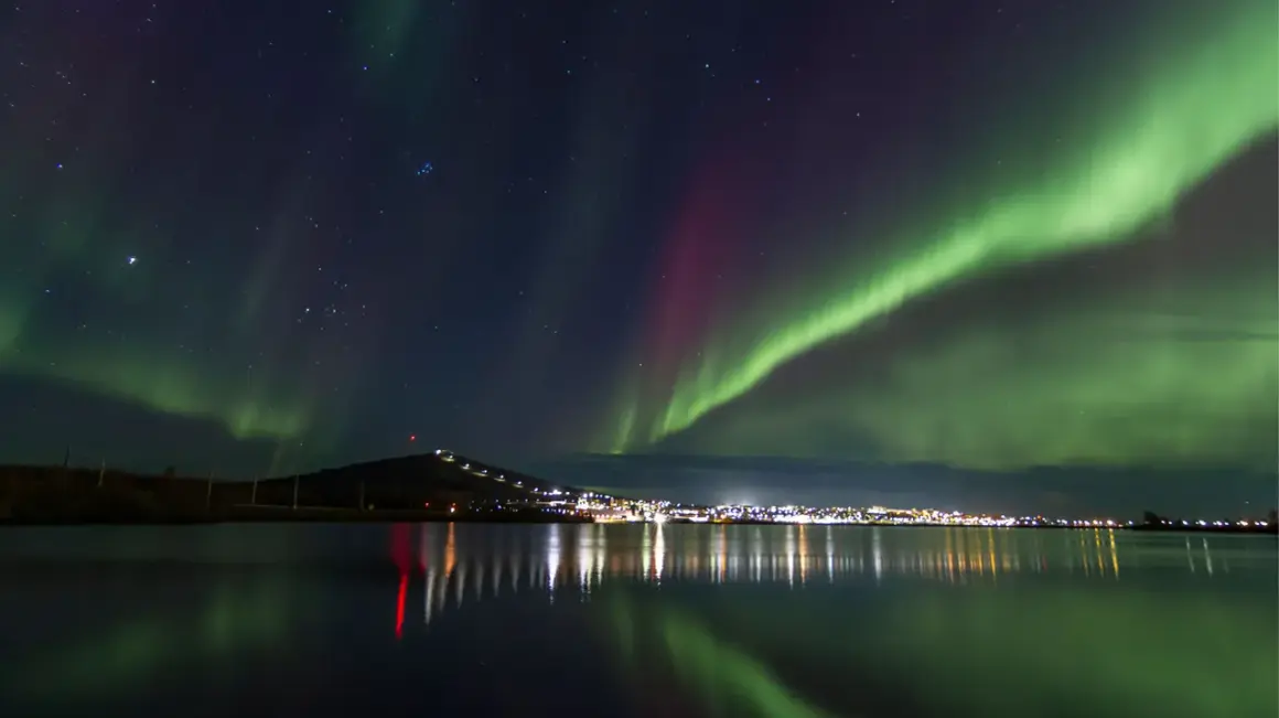 Kiruna norrsken lyser upp natthimlen över en stad vid en sjö. Gröna och lila ljusstrimmor dansar över stjärnorna.