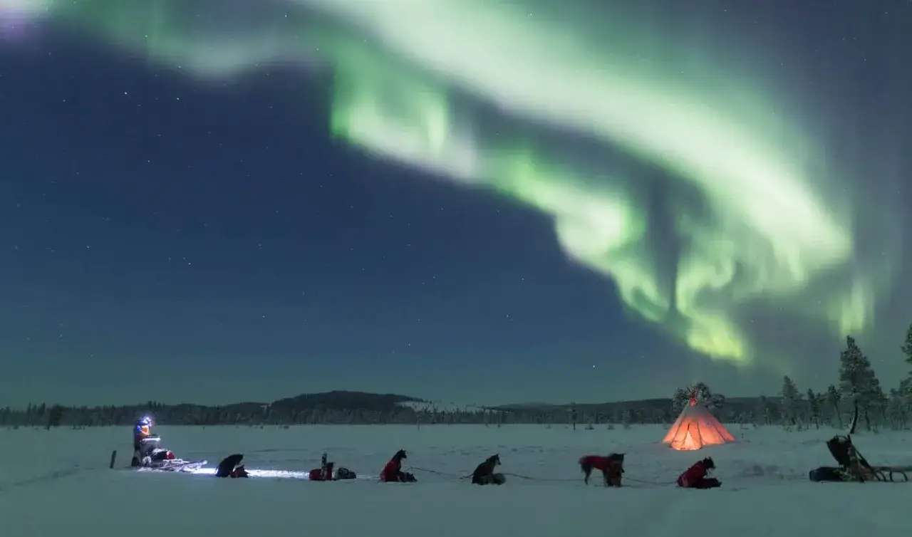 Magiskt kiruna norrsken &ouml;ver en sn&ouml;kl&auml;dd natt med hundspann och en upplyst k&aring;ta.