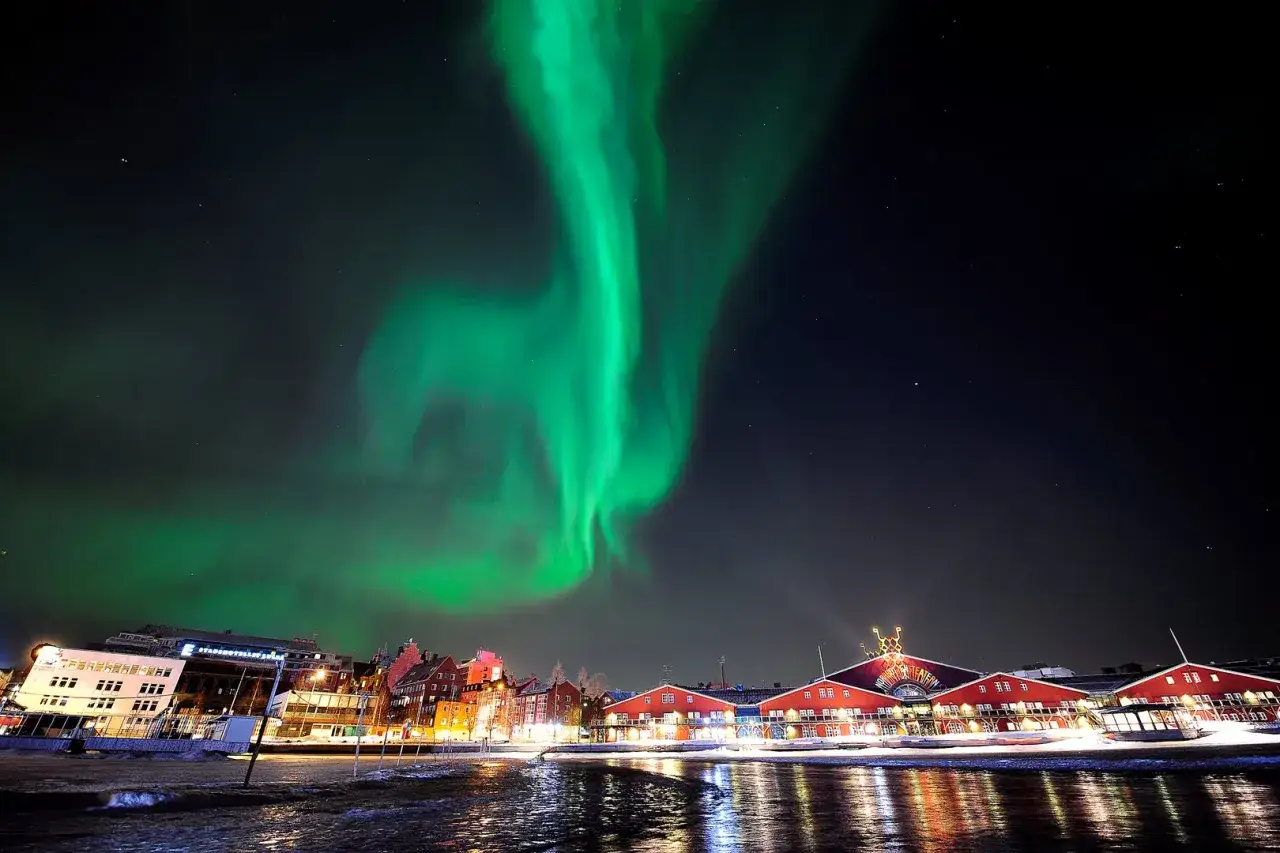 Grön norrskenshimmel över Luleå med röda byggnader och vatten som reflekterar ljusen.