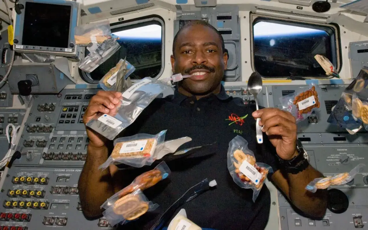 Astronaut njuter av rymdmat, h&aring;ller upp f&ouml;rpackningar med mat och en sked. En tub med v&auml;tska sitter i hans mun.