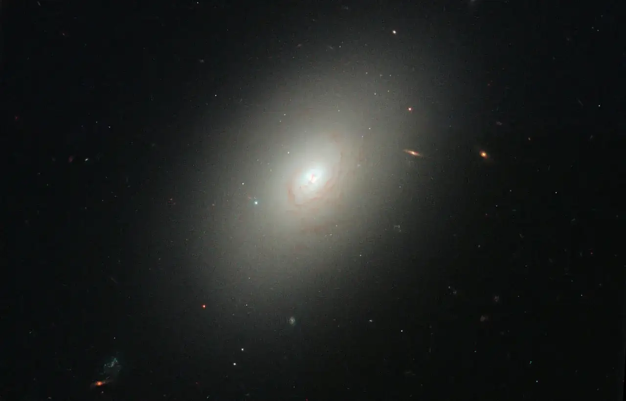 En stor, ljus elliptisk galax dominerar bilden, omgiven av mindre galaxer och stjärnor.
