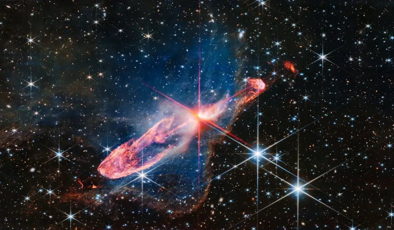 En lysande kometliknande nebulosa med en röd kärna, fotograferad av james webb-teleskopet, omgiven av stjärnor och galaxer.
