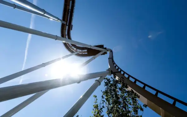 En berg-och-dalbana som svänger upp mot en klarblå himmel, solen lyser starkt och skapar en bländande effekt. Känslan av g krafter är påtaglig.