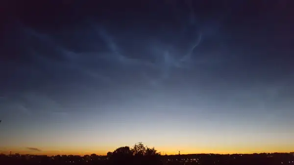 Mörka moln med ljusa strimmor, nattlysande moln, över en stad i skymningen.