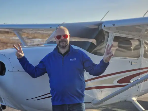 Man in blue shirt and red sunglasses making peace signs in front of a small airplane.